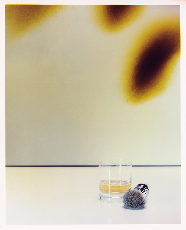 Still life photograph of a glass of whisky, a shaving brush and safety razor on a white surface, with a muted abstract background.