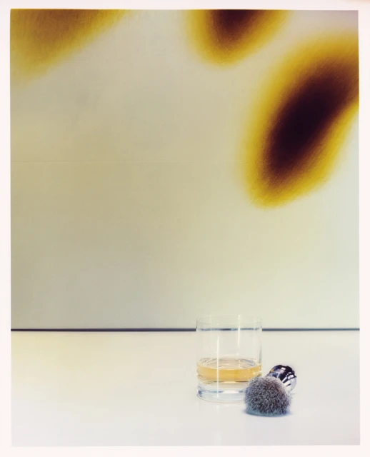 Still life photograph of a glass of whisky, a shaving brush and safety razor on a white surface, with a muted abstract background.