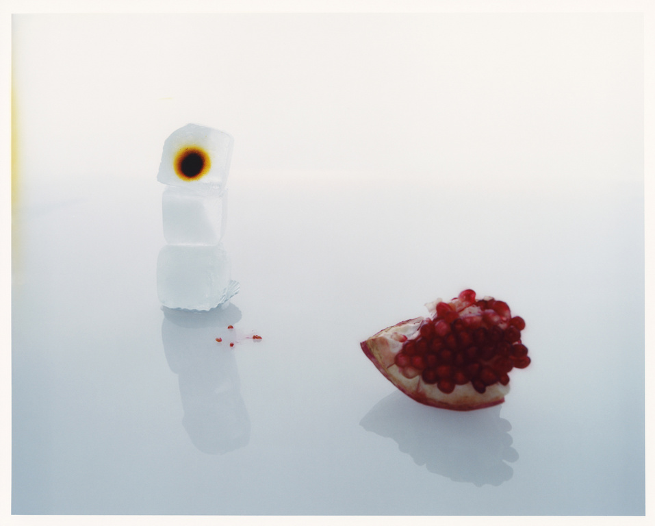 Still life photograph of a stack of three ice cubes with a dark brown stain and a halved pomegranate with seeds on a reflective surface.