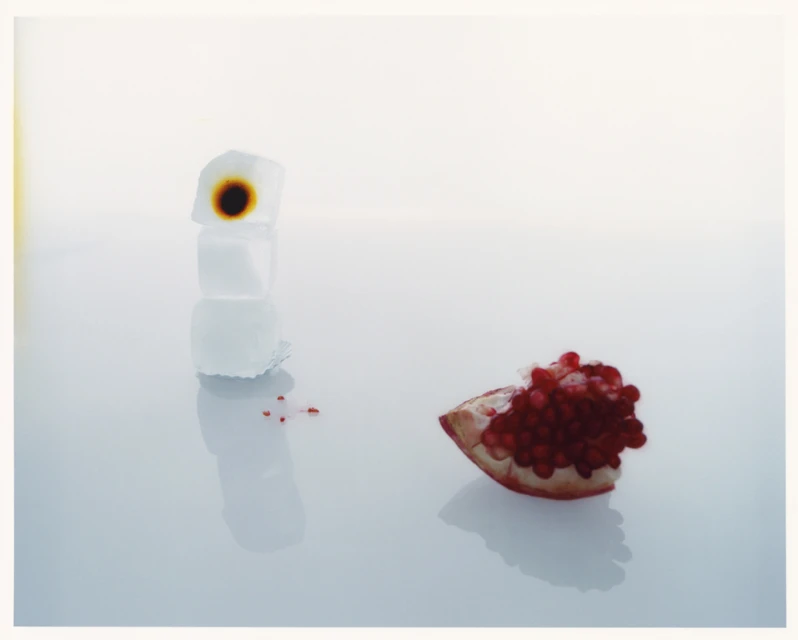 Still life photograph of a stack of three ice cubes with a dark brown stain and a halved pomegranate with seeds on a reflective surface.