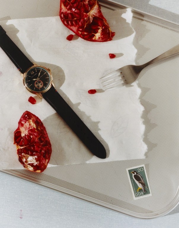 Wristwatch with black leather strap and gold casing displayed on a baking sheet with pomegranate halves and a fork, product shot.