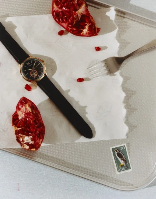 Wristwatch with black leather strap and gold casing displayed on a baking sheet with pomegranate halves and a fork, product shot.