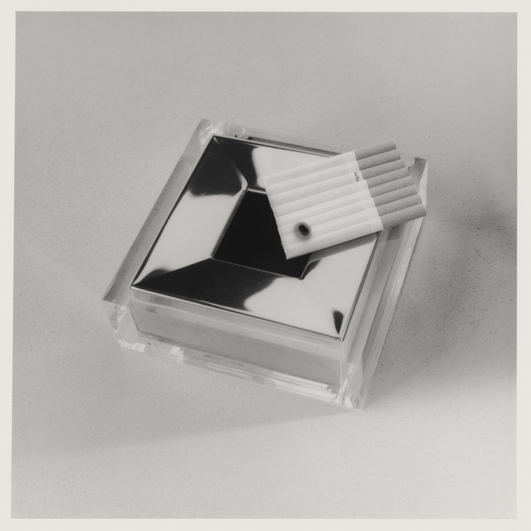 Close-up studio shot of a clear ashtray with a silver inset, accompanied by cigarettes in a plastic case, all on a neutral background.