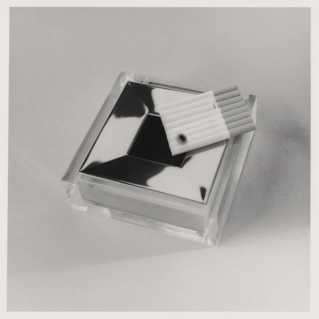 Close-up studio shot of a clear ashtray with a silver inset, accompanied by cigarettes in a plastic case, all on a neutral background.