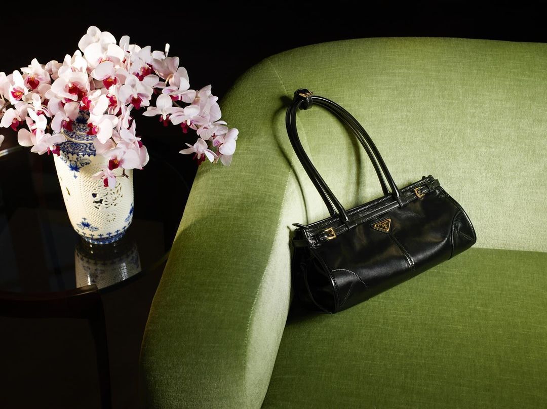 Black Prada purse on an apple green sofa with a blue and white vase holding white orchids on a dark glass table.