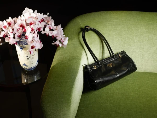 Black Prada purse on an apple green sofa with a blue and white vase holding white orchids on a dark glass table.