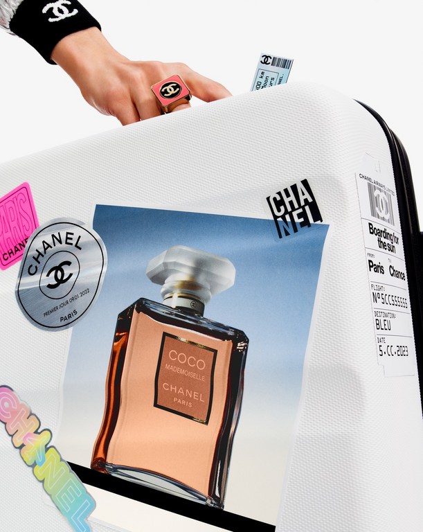 Close-up of a white suitcase with stickers and travel labels, featuring a Chanel perfume bottle image. Hand wearing Chanel accessories.