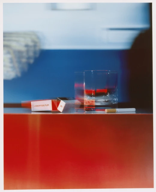 Still life shot on a red surface with a pack of cigarettes, a clear glass, and a single cigarette, set against a blue backdrop.
