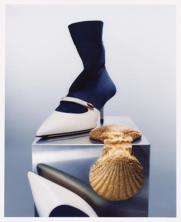 Still life shot of a white shoe with a blue sock-like upper on a reflective pedestal, with a gold seashell ornament nearby.