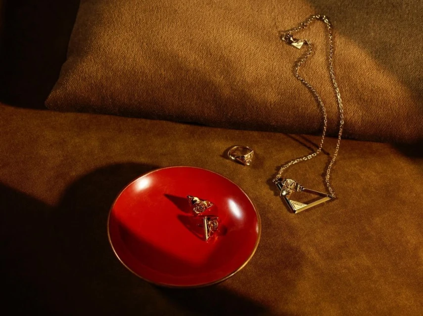 Close-up studio shot of gold jewelry including earrings, a ring, and necklace arranged on a red plate and a brown pillow.