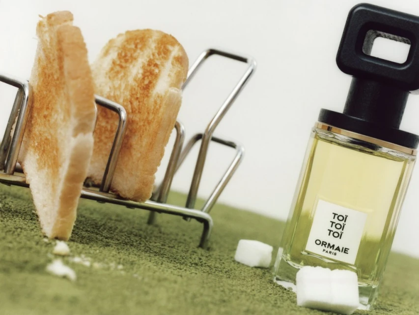 Still life featuring toasted bread in a wire rack, a rectangular perfume bottle, and sugar cubes on a green textured surface.