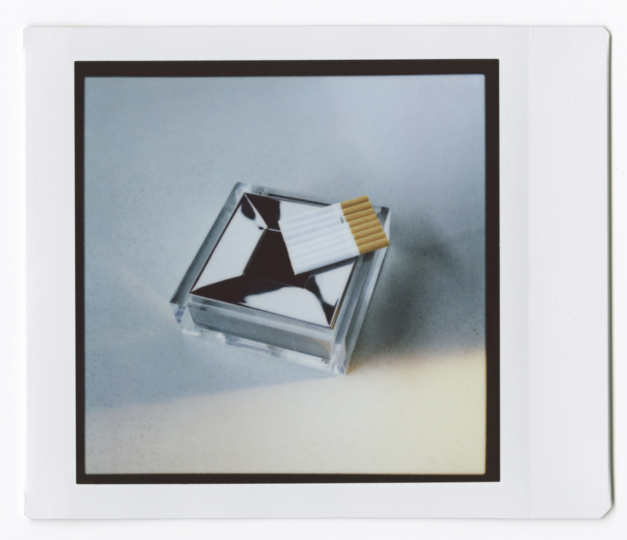 Still life photograph capturing cigarettes on top of a transparent ashtray with a modern design on a gradient grey background.