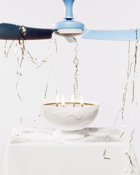 Lit candles sit in a white pedestal bowl below a light blue ceiling fan with silver tinsel on a white surface and background.