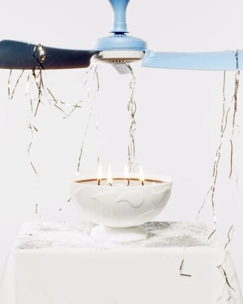 Lit candles sit in a white pedestal bowl below a light blue ceiling fan with silver tinsel on a white surface and background.