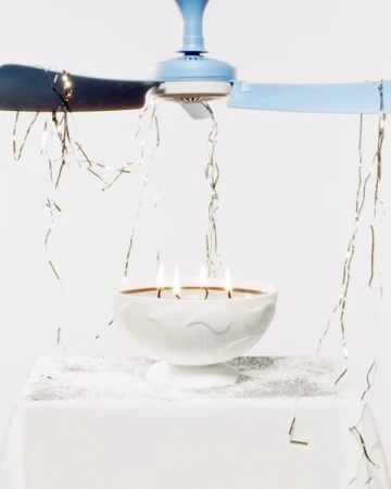 Lit candles sit in a white pedestal bowl below a light blue ceiling fan with silver tinsel on a white surface and background.