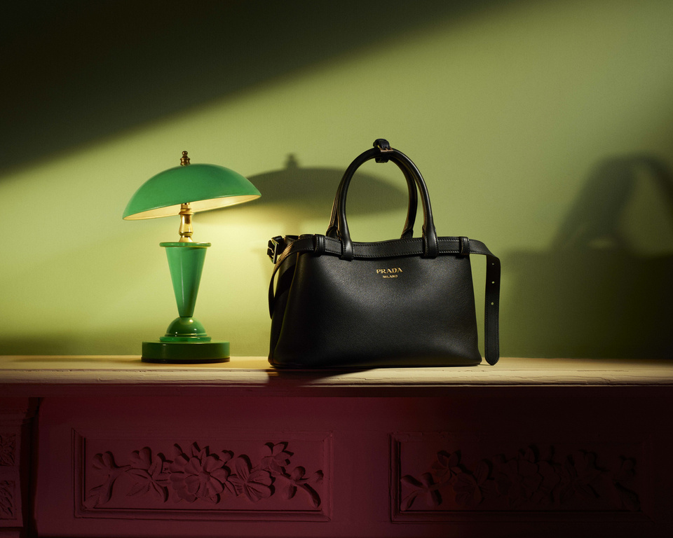 Black Prada handbag with gold logo and green desk lamp on wooden surface with decorative base against gradient green-red wall background.