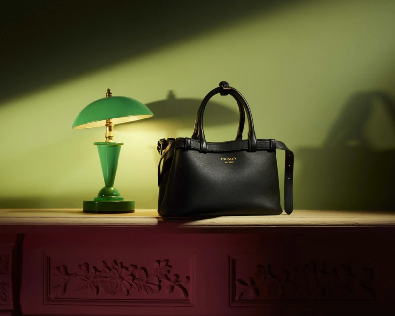 Black Prada handbag with gold logo and green desk lamp on wooden surface with decorative base against gradient green-red wall background.