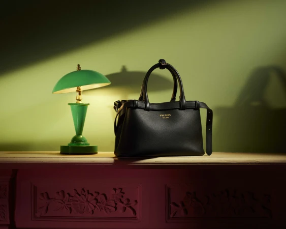 Black Prada handbag with gold logo and green desk lamp on wooden surface with decorative base against gradient green-red wall background.