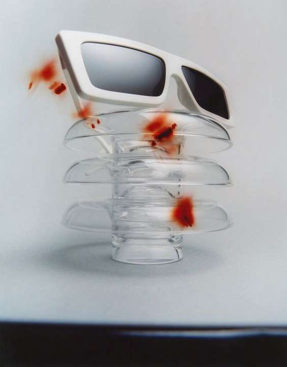 Fashion photography of white-rimmed sunglasses balanced atop a clear glass insulator, set against a gradated white and gray background.