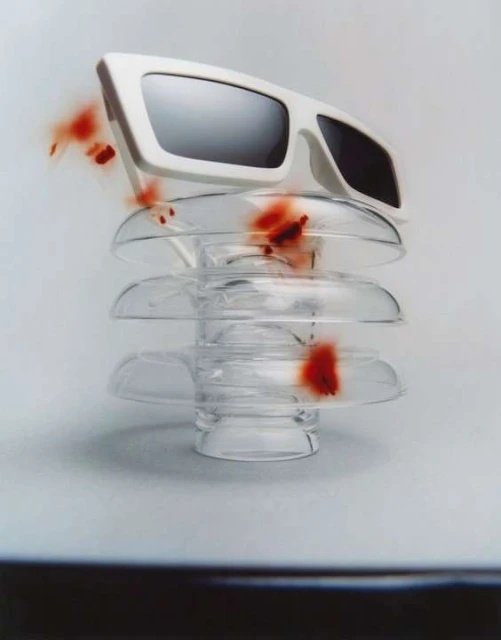 Fashion photography of white-rimmed sunglasses balanced atop a clear glass insulator, set against a gradated white and gray background.