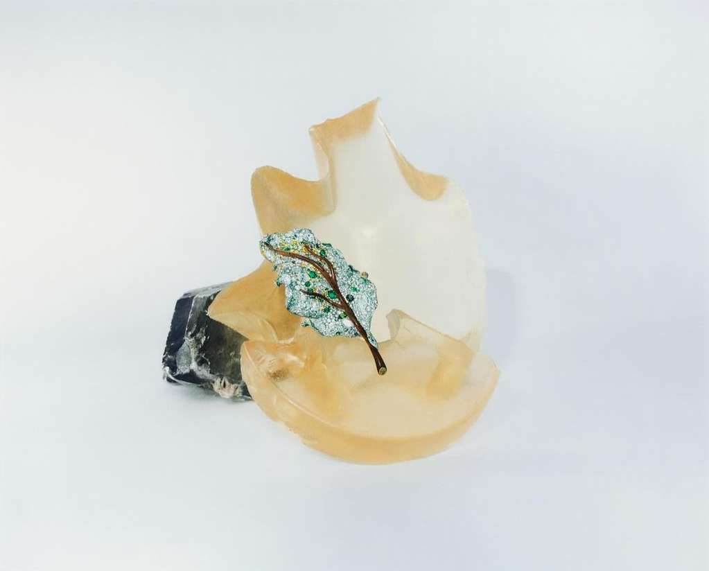 Still life of a green and silver ornamental leaf on a translucent form and grey rock against a clean white backdrop.