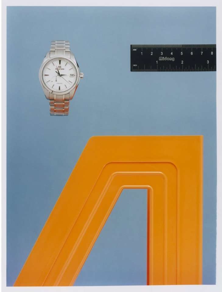Wristwatch on a bracelet next to a ruler and an orange geometric shape against a light blue background in a studio setting.