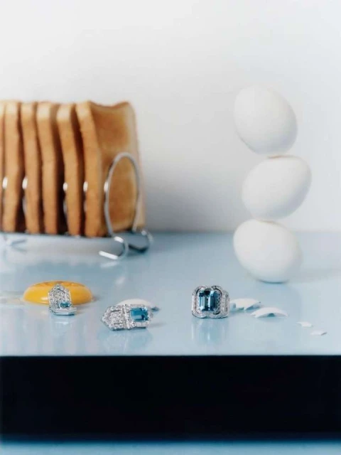 Still life photo of fine jewelry including rings and an earring with aquamarine stones set against a background of toast and eggs.