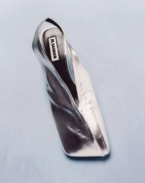 Elevated view of a metallic silver Jil Sander shoe on a light blue background, highlighting its sleek design and reflective surface.