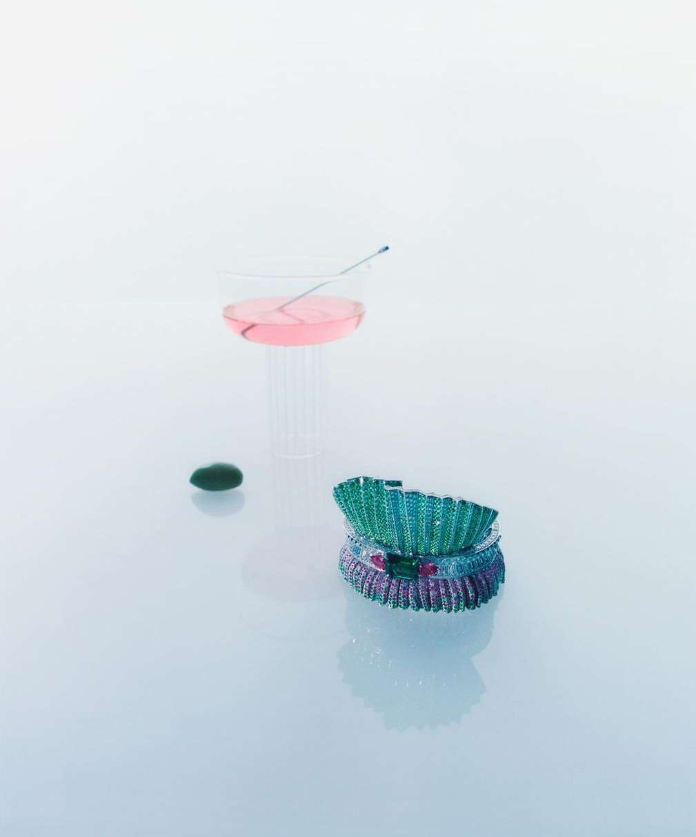Still life shot of stacked jewel-toned bracelets, a pink cocktail in a clear glass, and a green stone on a white reflective surface.