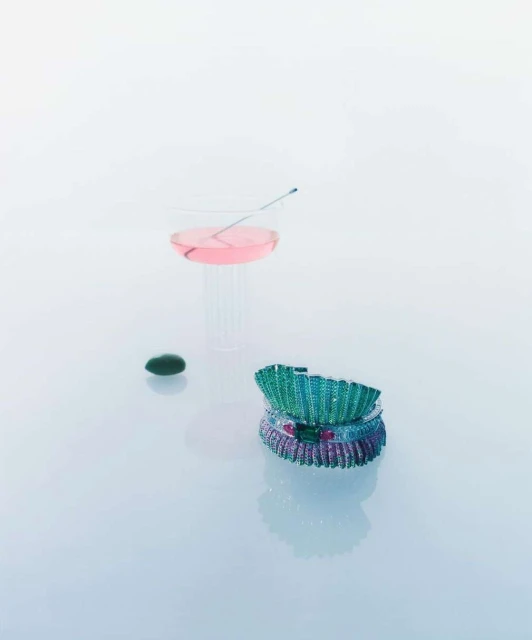 Still life shot of stacked jewel-toned bracelets, a pink cocktail in a clear glass, and a green stone on a white reflective surface.