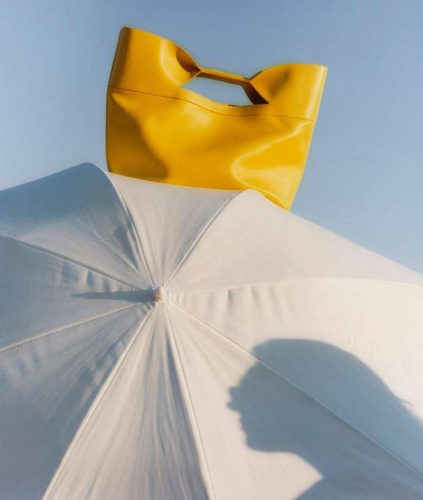 Bright yellow leather tote bag sits atop a white umbrella, contrasted against a light blue sky in this fashion product shot.