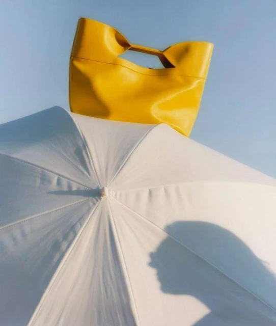Bright yellow leather tote bag sits atop a white umbrella, contrasted against a light blue sky in this fashion product shot.
