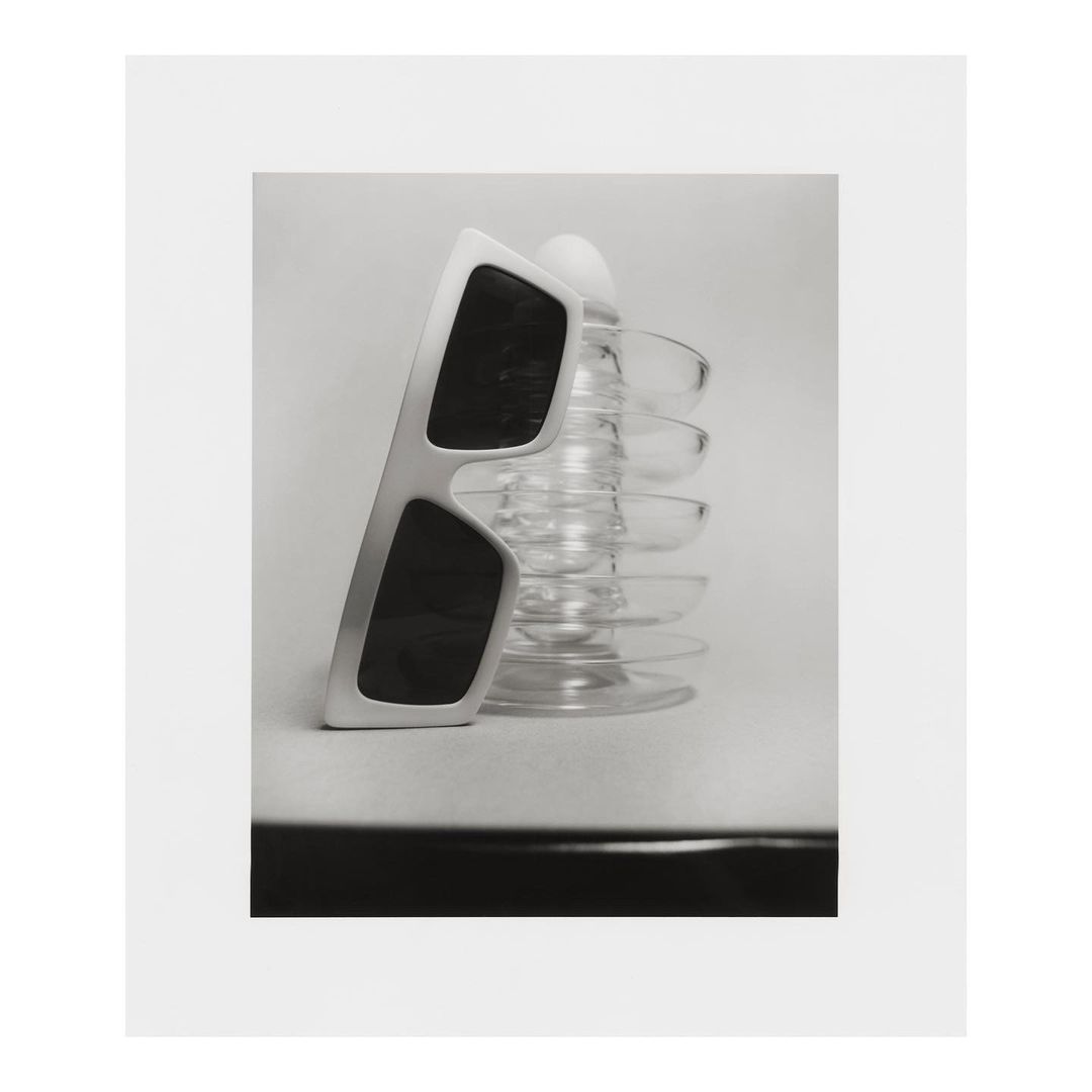 Still life photograph of sunglasses propped on a glass object, set against a clean, minimalist backdrop, offering a blend of shadow and light.
