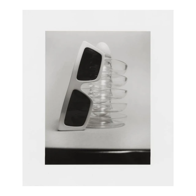 Still life photograph of sunglasses propped on a glass object, set against a clean, minimalist backdrop, offering a blend of shadow and light.
