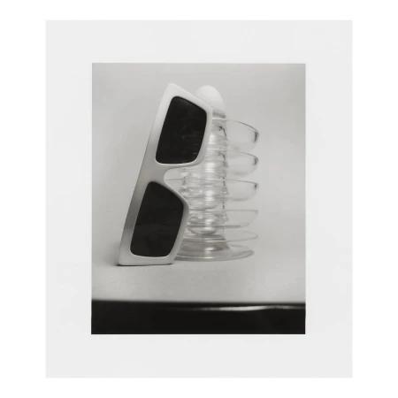 Still life photograph of sunglasses propped on a glass object, set against a clean, minimalist backdrop, offering a blend of shadow and light.