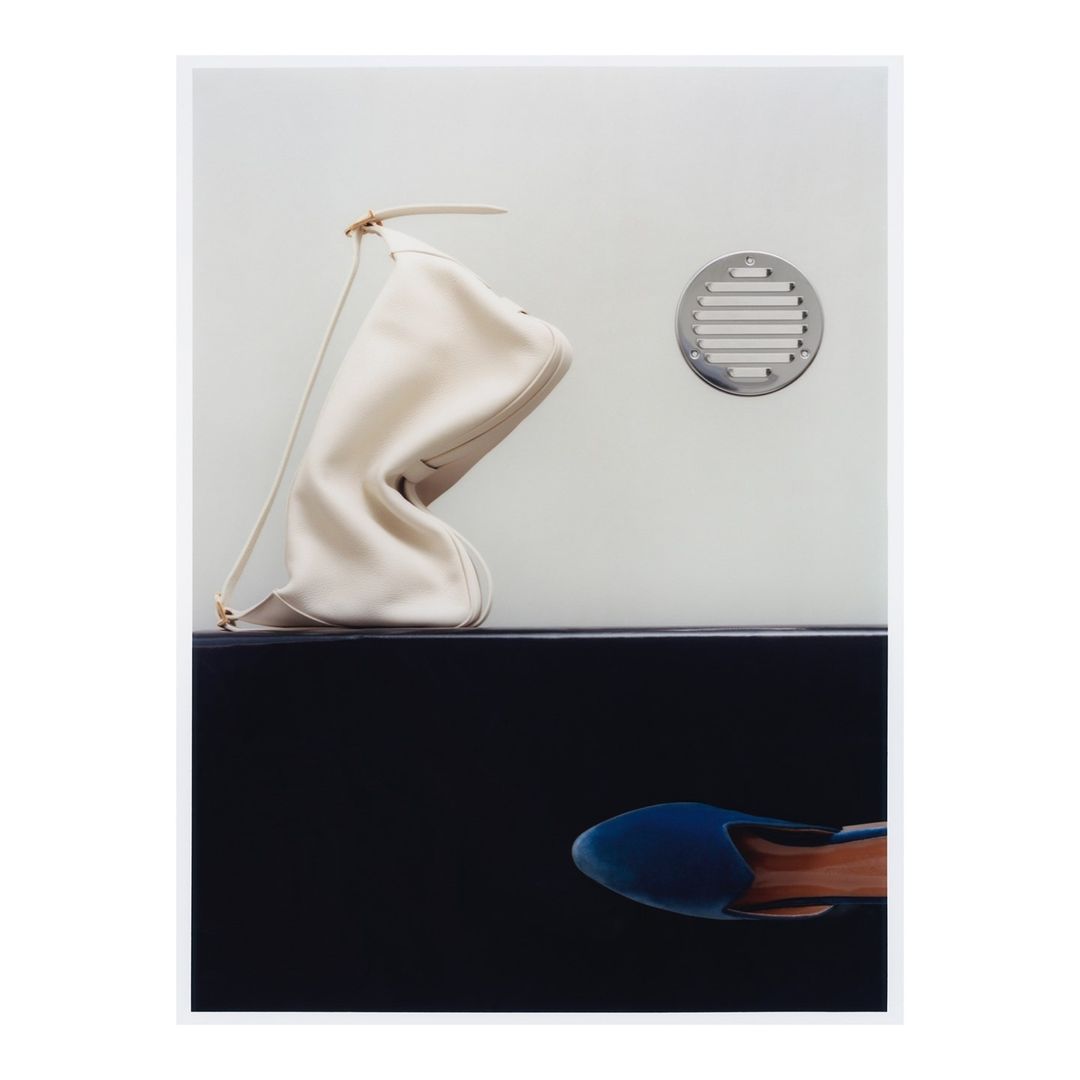 Fashion still life: Ivory purse on black shelf with a blue shoe beneath, set against a light grey wall with a metal vent.