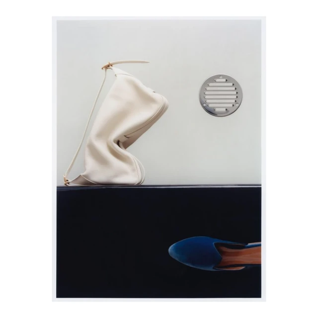 Fashion still life: Ivory purse on black shelf with a blue shoe beneath, set against a light grey wall with a metal vent.
