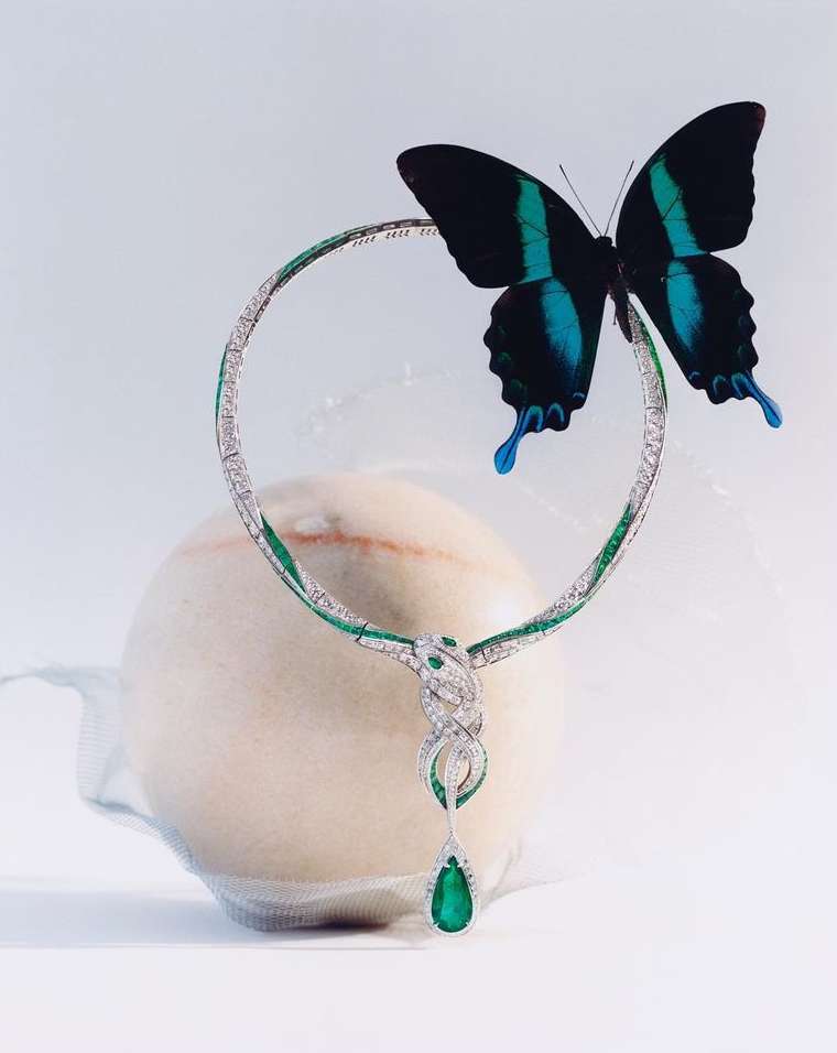 Necklace with diamonds and emerald, adorned by butterfly with blue markings, rests on a blurry round surface against a light backdrop.