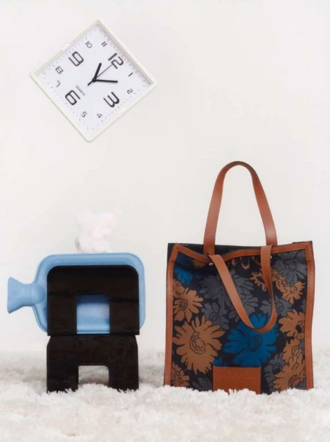 Floral tote bag stands next to a blue container on a stack of black cushions; square white clock on a white wall above the scene.