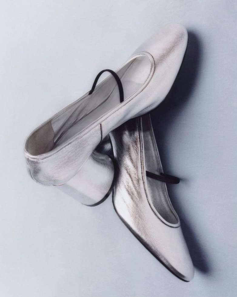 Shiny silver Mary Jane shoes with a black strap, displayed on a light blue background for a fashion-forward studio product shot.