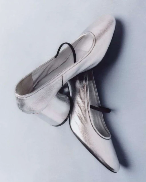 Shiny silver Mary Jane shoes with a black strap, displayed on a light blue background for a fashion-forward studio product shot.