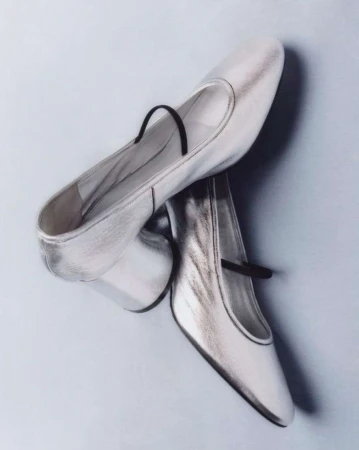 Shiny silver Mary Jane shoes with a black strap, displayed on a light blue background for a fashion-forward studio product shot.