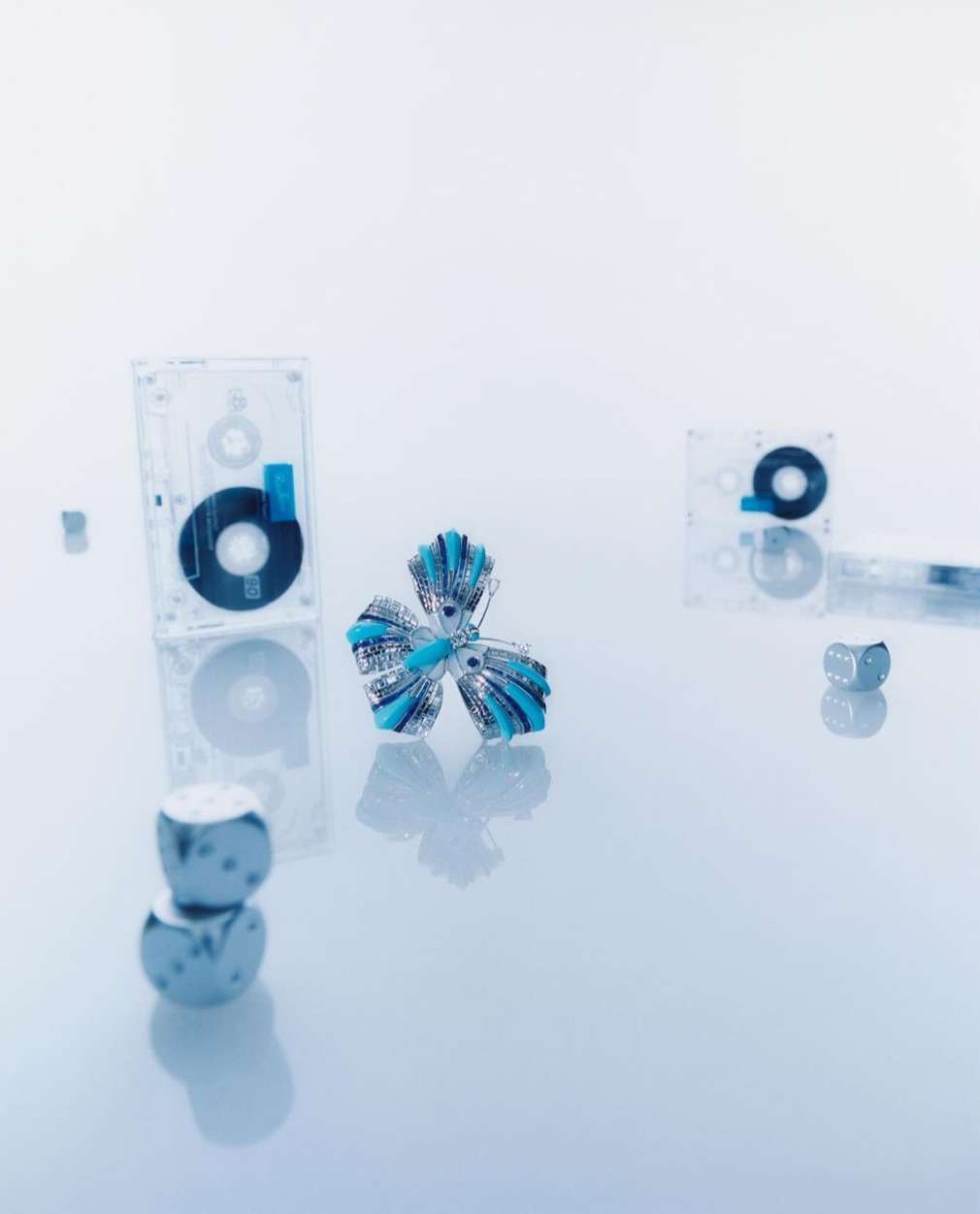 Modern product shot of teal and silver flower surrounded by silver cubes and retro cassette tapes on a white reflective surface.