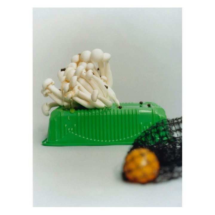 Still life shot of white beech mushrooms on a green plastic container with a black net and a yellow fruit against a neutral background.