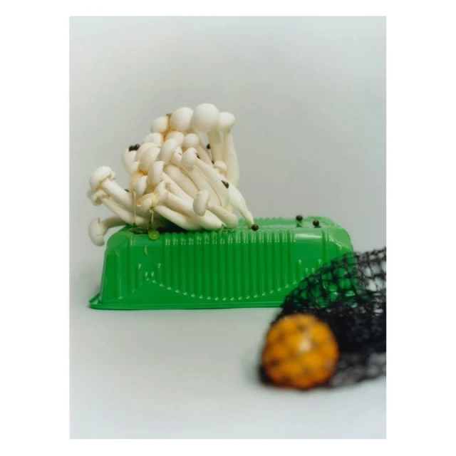 Still life shot of white beech mushrooms on a green plastic container with a black net and a yellow fruit against a neutral background.