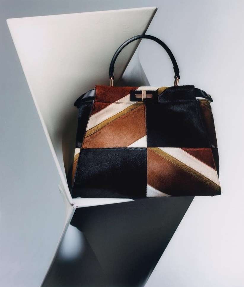 Geometric patchwork leather and fur handbag with black handle, featuring black, brown, and white tones, staged against a minimalist white backdrop.
