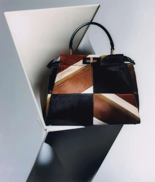 Geometric patchwork leather and fur handbag with black handle, featuring black, brown, and white tones, staged against a minimalist white backdrop.