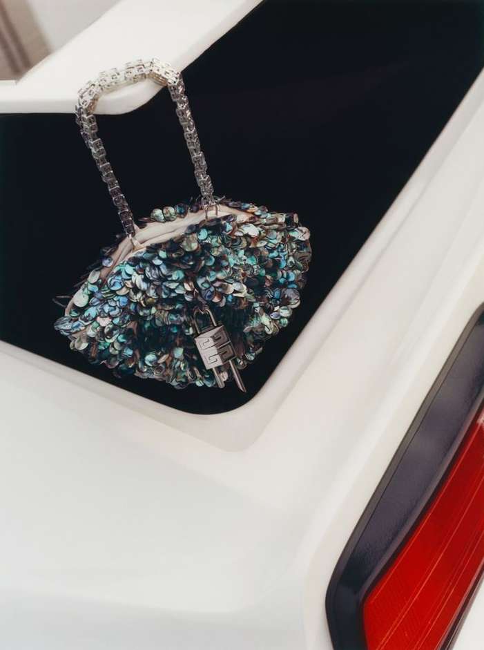Close-up of a shimmering beaded clutch handbag with a chain handle displayed inside of the open trunk of a glossy white vehicle.