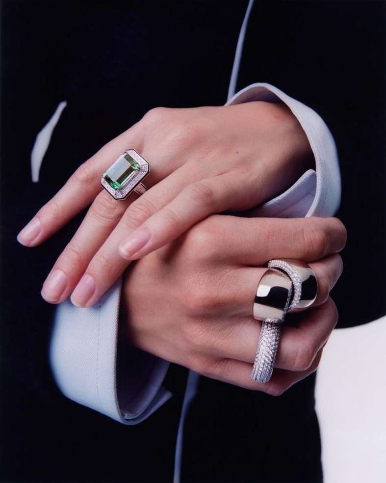 Close-up of an adult female's crossed hands showcasing rings with emerald-cut green gemstones and diamond accents set against a black suit.