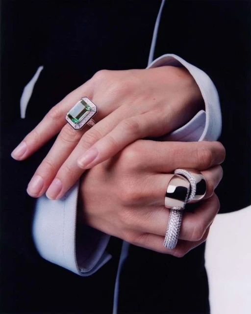 Close-up of an adult female's crossed hands showcasing rings with emerald-cut green gemstones and diamond accents set against a black suit.